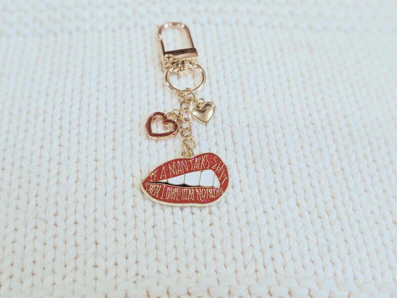 May include: Gold-tone keychain with a red lips charm that reads "IF A MAN TALKS SHIT THEN LOVE HIM NOTHIN". The keychain also features two heart charms. The background is a white, textured surface.
