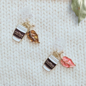 May include: A pair of coffee cup earrings with gold-colored accents and leaf charms. The coffee cups are white with black trim and the word "coffee" printed on them. One leaf is gold, the other is red.
