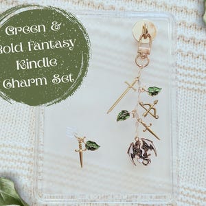 Op de afbeelding: Een goudkleurige bedelset met fantasiethema-hangers, waaronder zwaarden, een pijl en boog en een draak. De bedels zijn geaccentueerd met groene bladeren. De set wordt weergegeven op een transparante achtergrond met de tekst "Green & Gold Fantasy Kindle Charm Set".