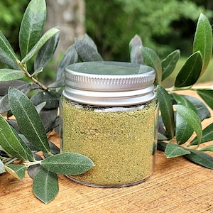 May include: A small, clear glass jar with a silver lid filled with a fine, green powder. The jar is surrounded by green olive branches and sits on a wooden surface. The background is blurred, suggesting an outdoor setting.