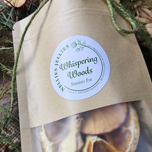 May include: A brown paper pouch containing dried fruit slices, tied with green twine. The label reads Nellies Jellies Whispering Woods Simmer Pot, with a note stating it is not produced in a regulated or licensed facility.