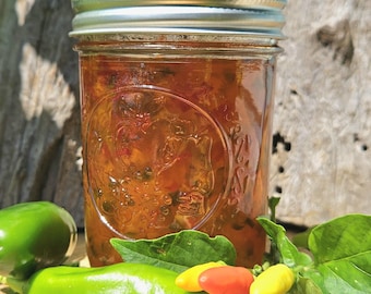 Pode incluir: Um frasco de vidro cheio de geleia de pimenta laranja e vermelha brilhante. O frasco tem uma tampa prateada e está sentado em uma superfície de madeira. Há pimentas verdes e vermelhas em primeiro plano.