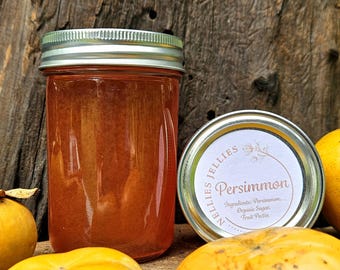 May include: A jar of persimmon jelly with a silver lid, next to a jar lid with the word "Persimmon" and the ingredients listed. The jelly is a reddish-orange color. Several ripe persimmons are in the foreground.