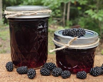 May include: Two glass jars filled with dark purple liquid, likely jam or jelly, with silver lids and twine accents. Fresh blackberries are scattered around the jars on a wooden surface, suggesting homemade preserves.
