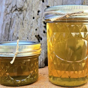 May include: Two glass jars filled with a golden liquid, likely a preserve or honey, sit next to two orange pumpkins. The jars have silver lids and are decorated with twine. The background features a weathered tree trunk.
