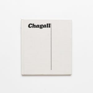 May include: A square, off-white book with the title "Chagall" in bold, black font. A vertical black line extends from the bottom of the title. The book has a slightly textured surface and is set against a white background.