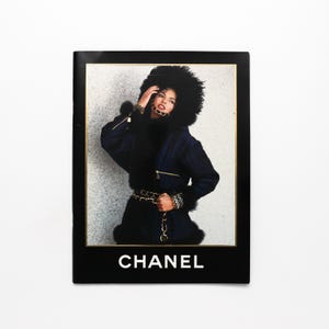 Könnte beinhalten: Modeanzeige mit einer Frau in einem dunkelblauen Mantel mit Kapuze und Manschetten mit Pelzbesatz. Der Mantel hat goldene Reißverschlüsse und einen goldenen Kettenriemen. Das Wort "CHANEL" ist in Weiß gedruckt.