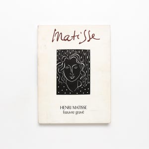 Puede incluir: Un libro titulado "Matisse" con un retrato en blanco y negro de una mujer en la portada. El título está escrito en una escritura de color rojo oscuro encima del retrato. El libro también incluye el texto "HENRI MATISSE l'oeuvre gravé".