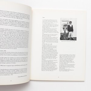 K&ouml;nnte beinhalten: Ein Schwarzwei&szlig;foto eines Mannes, der vor einem gro&szlig;en, farbenfrohen Gem&auml;lde steht. Das Gem&auml;lde zeigt ein gro&szlig;es, abstraktes Gesicht mit einem roten Mund und einem blauen Auge. Der Mann tr&auml;gt ein wei&szlig;es Hemd und eine schwarze Hose. Das Gem&auml;lde ist mit "Philosophie, Jean-Michel Basquiat, 1982" betitelt.