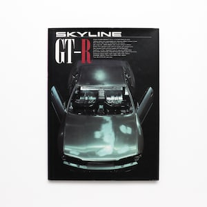 Peut inclure: Une couverture de magazine présentant une voiture argentée avec les portes ouvertes. Le texte "SKYLINE" est en haut, avec "GT-R" en grandes lettres en dessous. L'intérieur de la voiture est visible, et le fond est sombre.