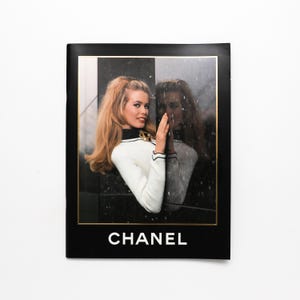Könnte beinhalten: Vintage Chanel-Werbung mit einer Frau in einem weiß-schwarzen Pullover und Goldschmuck. Das Bild ist schwarz umrahmt und trägt unten in Weiß den Markennamen "CHANEL".