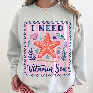 May include: A light gray crewneck sweatshirt featuring a central graphic with pink vertical stripes and a dark blue wave border. The design includes a large orange starfish, seashells, seahorses, and jellyfish with the text I NEED Vitamin Sea.