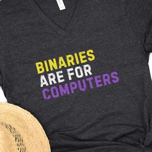 May include: Dark gray v-neck t-shirt with the text "BINARIES ARE FOR COMPUTERS" in yellow, white, and purple. The shirt is made of a soft material and is suitable for casual wear. A straw hat is partially visible in the image.