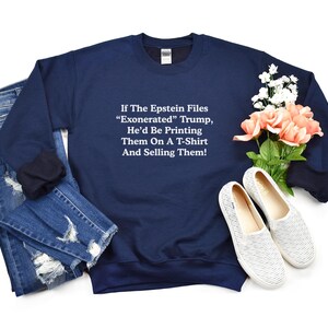May include: Navy blue sweatshirt with white text: "If The Epstein Files 'Exonerated' Trump, He'd Be Printing Them On A T-Shirt And Selling Them!" The sweatshirt is paired with blue jeans and white shoes with a floral arrangement.
