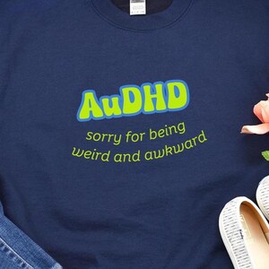 May include: Navy blue sweatshirt with the text "AuDHD" in bright green and blue, with the phrase "sorry for being weird and awkward" below. The sweatshirt is a casual style.