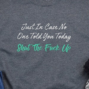 May include: Dark gray t-shirt with white text that reads "Just In Case No One Told You Today Shut The Fuck Up". The text "Shut The Fuck Up" is in a teal green color.