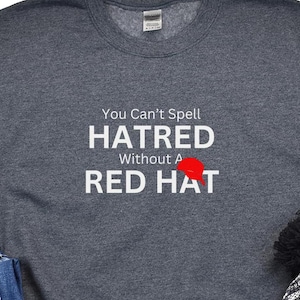 May include: A dark gray sweatshirt with the text "You Can't Spell HATRED Without A RED HAT" in white letters. A red baseball cap is shown in the text.