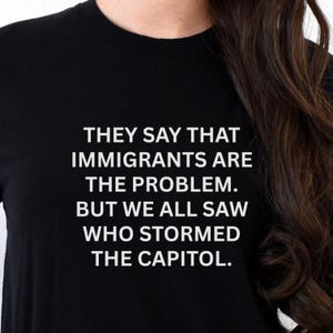 May include: Black t-shirt with white text. The text reads: "THEY SAY THAT IMMIGRANTS ARE THE PROBLEM. BUT WE ALL SAW WHO STORMED THE CAPITOL."