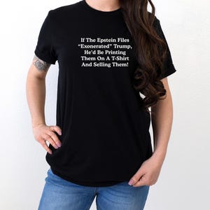 May include: Black t-shirt with white text: "If The Epstein Files 'Exonerated' Trump, He'd Be Printing Them On A T-Shirt And Selling Them!" The shirt is worn by a person in blue jeans.