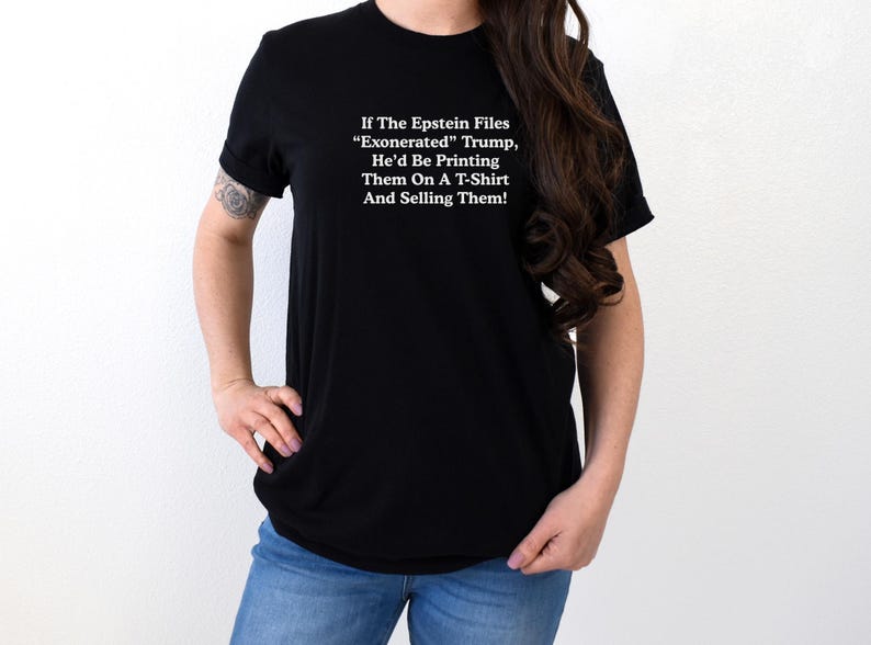 May include: Black t-shirt with white text: "If The Epstein Files 'Exonerated' Trump, He'd Be Printing Them On A T-Shirt And Selling Them!" The shirt is worn by a person in blue jeans.