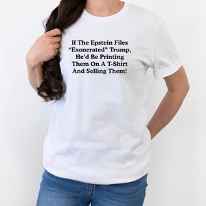 May include: White t-shirt with black text. The text reads: "If The Epstein Files 'Exonerated' Trump, He'd Be Printing Them On A T-Shirt And Selling Them!" The shirt is worn by a person wearing blue jeans.