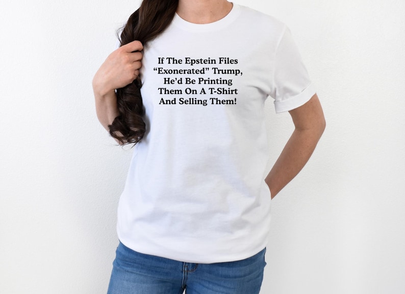 May include: White t-shirt with black text. The text reads: "If The Epstein Files 'Exonerated' Trump, He'd Be Printing Them On A T-Shirt And Selling Them!" The shirt is worn by a person wearing blue jeans.