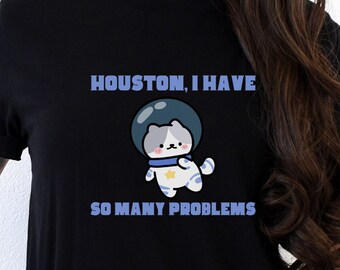 May include: Black t-shirt with a white cartoon cat wearing a blue astronaut helmet. The text "HOUSTON, I HAVE SO MANY PROBLEMS" is printed on the shirt.
