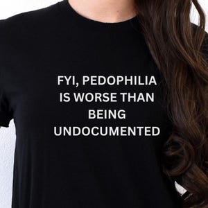 May include: Black t-shirt with white text that reads "FYI, PEDOPHILIA IS WORSE THAN BEING UNDOCUMENTED". The shirt is made of a dark fabric and has a crew neck.