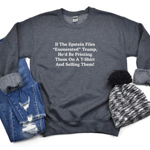 May include: A dark gray sweatshirt with the text "If The Epstein Files 'Exonerated' Trump, He'd Be Printing Them On A T-Shirt And Selling Them!" is displayed with a pair of blue jeans and a black and white beanie.