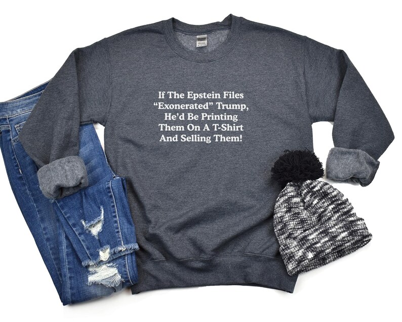 May include: A dark gray sweatshirt with the text "If The Epstein Files 'Exonerated' Trump, He'd Be Printing Them On A T-Shirt And Selling Them!" is displayed with a pair of blue jeans and a black and white beanie.