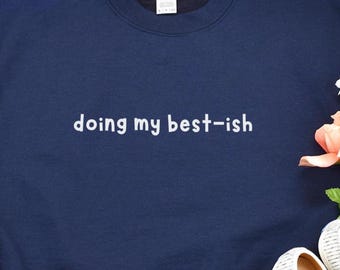 May include: Navy blue sweatshirt with the phrase "doing my best-ish" in white lettering. The sweatshirt has a crew neck and long sleeves. The image also shows a pair of white shoes and a pink flower.
