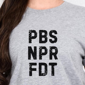 May include: A heather gray t-shirt with the black text "PBS NPR FDT" printed on the front. The letters have a distressed, textured appearance. The shirt is made of a soft material.