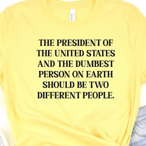 May include: Yellow t-shirt with black text. The text reads: "THE PRESIDENT OF THE UNITED STATES AND THE DUMBEST PERSON ON EARTH SHOULD BE TWO DIFFERENT PEOPLE."