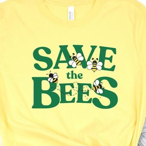 May include: Yellow t-shirt with the text "SAVE the BEES" in green block letters. The word "SAVE" has three cartoon bees, and the word "BEES" has two cartoon bees. The bees are yellow, black, and white.