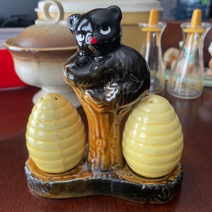 May include: A ceramic salt and pepper shaker set featuring a black bear figurine perched on a tree stump. The shakers are shaped like honeycombs and are a pale yellow colour.