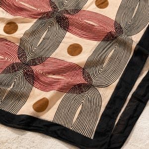 May include: A lightweight fabric scarf featuring a cream-colored base with a repeating pattern of black and dark red concentric oval and arch shapes, interspersed with solid brown circles, finished with a solid black border along the edge.