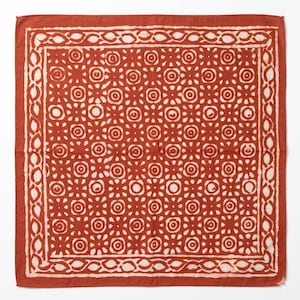 Pode incluir: Uma bandana quadrada, cor de ferrugem, com um padrão floral branco repetido. A bandana tem uma borda branca com um design geométrico. O tecido parece ser algodão ou material semelhante.