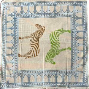 May include: A white cotton square scarf with a blue border and a pattern of two zebras, one brown and one green, on a pink background.