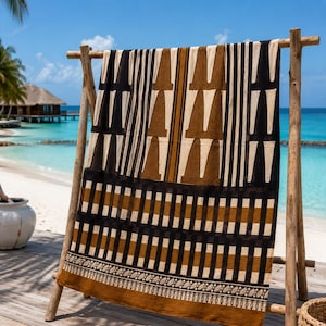 Puede incluir: Un textil rectangular colgado en un marco de madera, con un fondo crema y patrones geom&eacute;tricos en negro y marr&oacute;n. El dise&ntilde;o incluye rayas verticales, formas triangulares apiladas, un borde a cuadros y un dobladillo con un patr&oacute;n de hojas detallado.