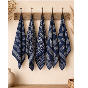 May include: Five navy blue patterned kitchen towels hanging on a wooden rack with metal hooks. Each towel features a unique white geometric design. The towels are displayed against a neutral wall, with a vase and basket in the background.