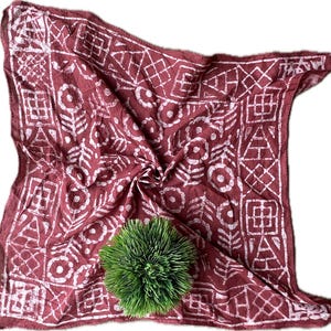 May include: A red and white patterned cotton bandana with a white geometric design. The bandana is folded and has a green plant in the center.