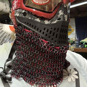 May include: A red and black paintball mask with a skull and crossbones pattern. The mask features a clear lens with the text "THERMAL FOG RESISTANT LENS". A chainmail neck protector with red and silver rings is attached.