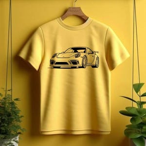 May include: Yellow t-shirt featuring a black graphic of a sports car. The car design includes details like headlights, wheels, and a spoiler. The shirt is hanging on a wooden hanger against a yellow background.