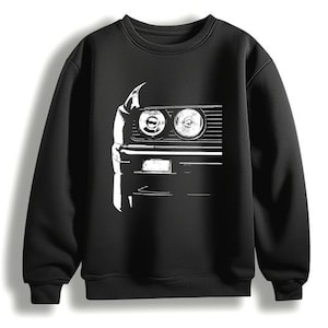 Pode incluir: Camisola preta de gola redonda com um gráfico branco que representa a frente de um carro, incluindo faróis, grelha e pára-choques. O design é uma ilustração minimalista em dois tons. A camisola é feita de material macio.