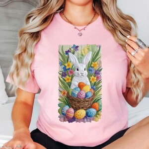 Puede incluir: Camiseta rosa claro con un diseño de un conejo blanco rodeado de flores coloridas y una cesta de huevos de Pascua. El diseño es de estilo acuarela, con colores vibrantes y un tema primaveral.