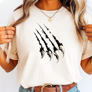 Puede incluir: Camiseta color crema con un diseño de garra negra con detalles blancos. Las garras están en ángulo diagonalmente a través de la camiseta. La camiseta está hecha de un material suave.
