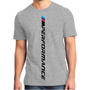 Op de afbeelding: Een grijs t-shirt met een verticaal ontwerp. Het ontwerp heeft een blauw, rood en witte streep bovenaan, gevolgd door het woord "IMPERFORMANCE" in het zwart, verticaal uitgelijnd.