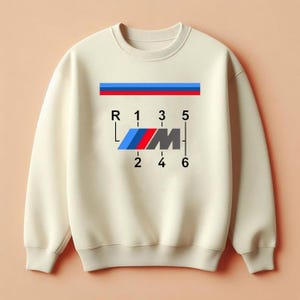 May include: A cream-coloured sweatshirt with a car gear shift pattern graphic. The design incorporates a blue, red, and black logo, along with the numbers and letters R, 1, 2, 3, 4, 5, and 6. A blue and red stripe is at the top of the design.