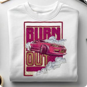 May include: White crewneck sweatshirt featuring a pink sports car design with smoke and the words "BURN OUT" in a pink and gold embroidered style. The car is in motion, creating a dynamic visual.