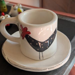 Può includere: Un set di tazza e piattino da espresso in ceramica bianca. La tazza presenta un design nero con pois bianchi e un fiore rosso. Il piattino ha un design abbinato. Il set è su una superficie scura.